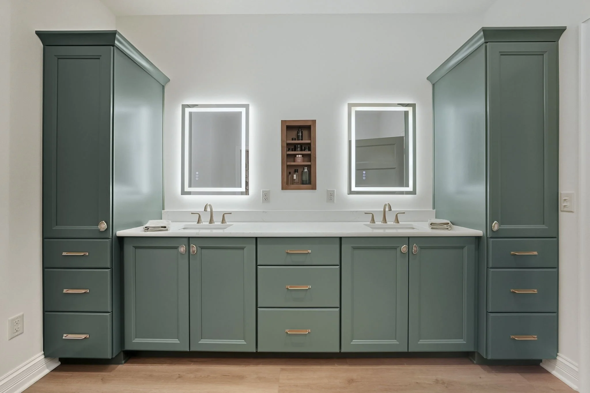 Double vanity bathroom with green cabinets, two rectangular illuminated mirrors, small wall shelf with toiletries, and two sinks with gold fixtures.