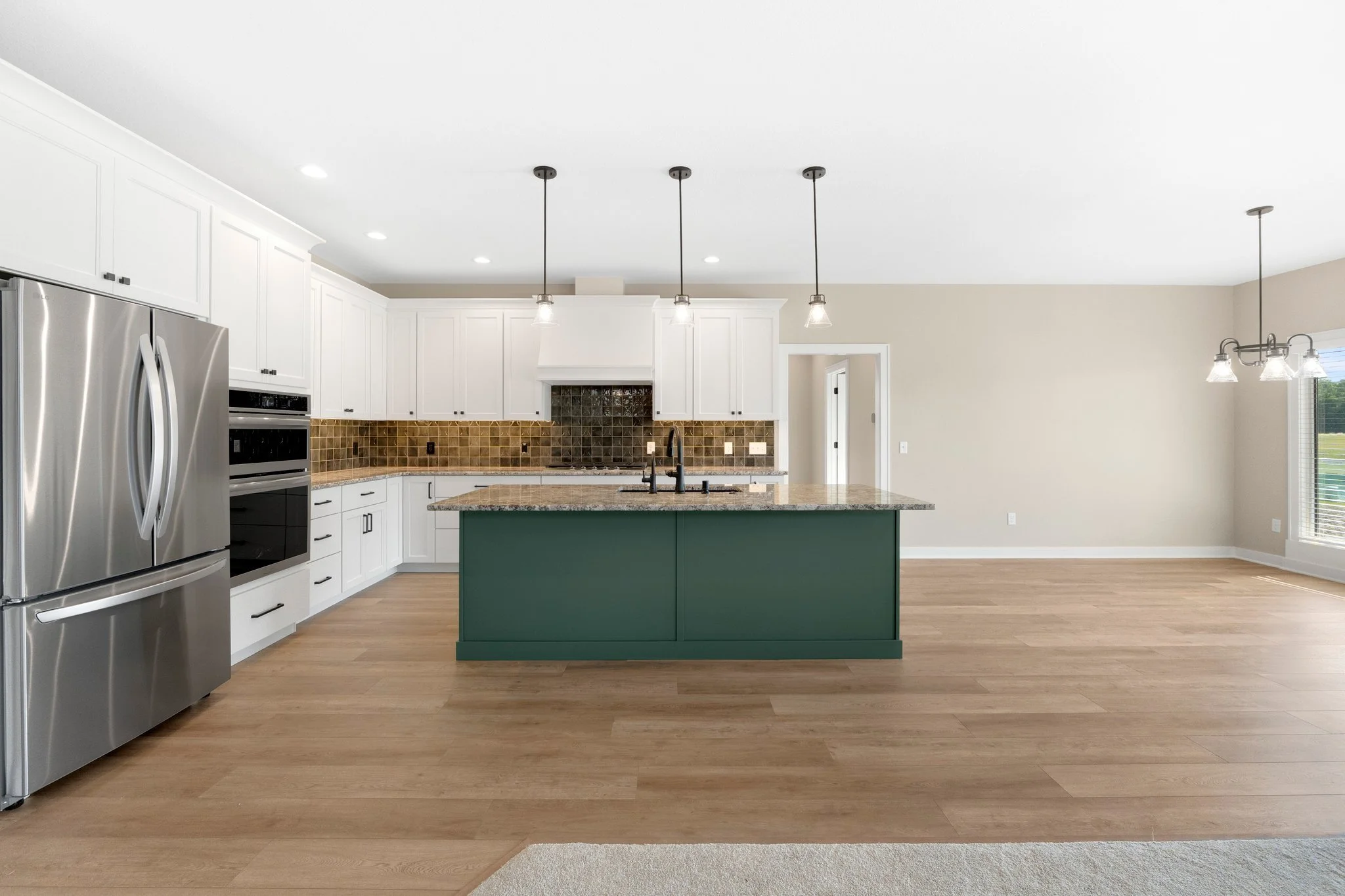 Modern kitchen with white cabinets, stainless steel refrigerator, granite countertops, a green island with a sink, brown tiled backsplash, and pendant lighting, with hardwood flooring and a spacious open layout.