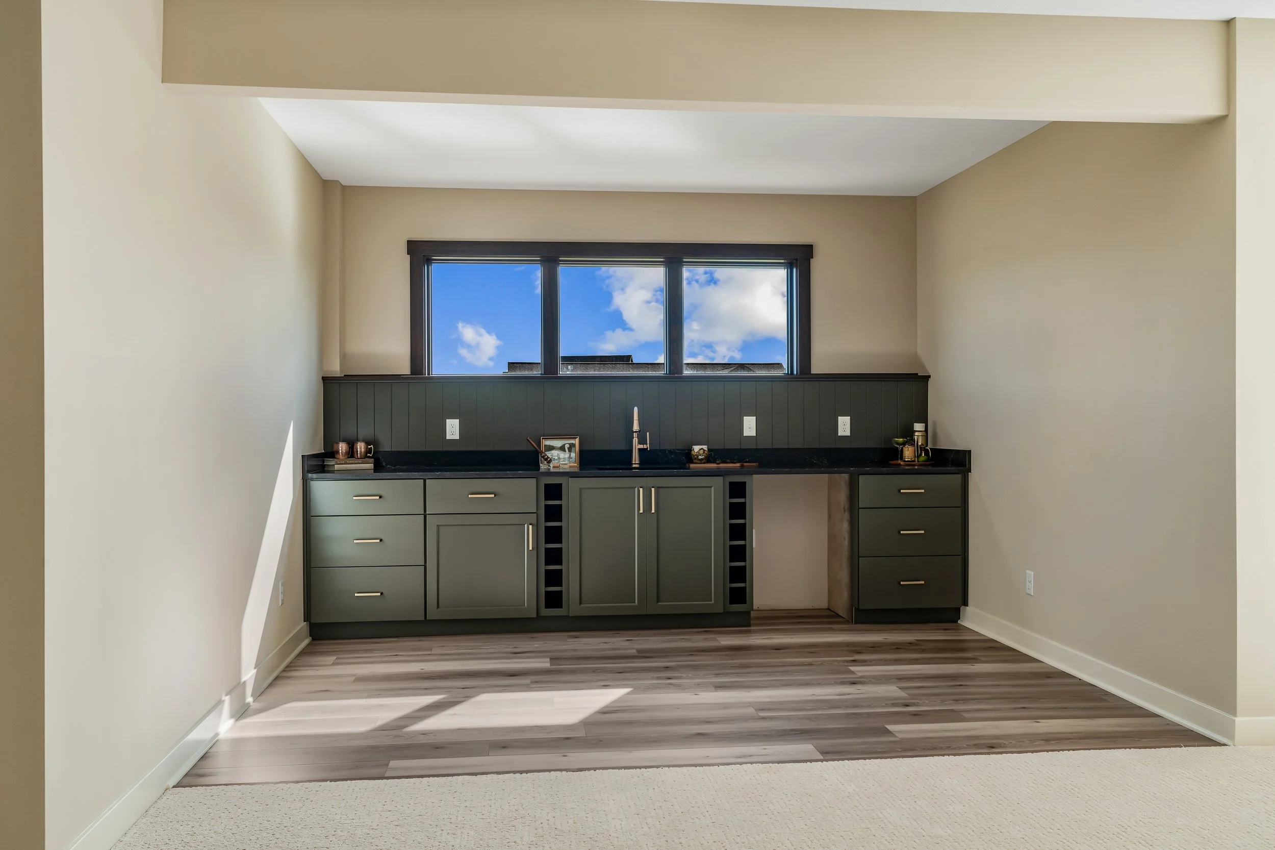 Empty kitchen with dark gray cabinets, black countertops, and a large window showing a blue sky with clouds.