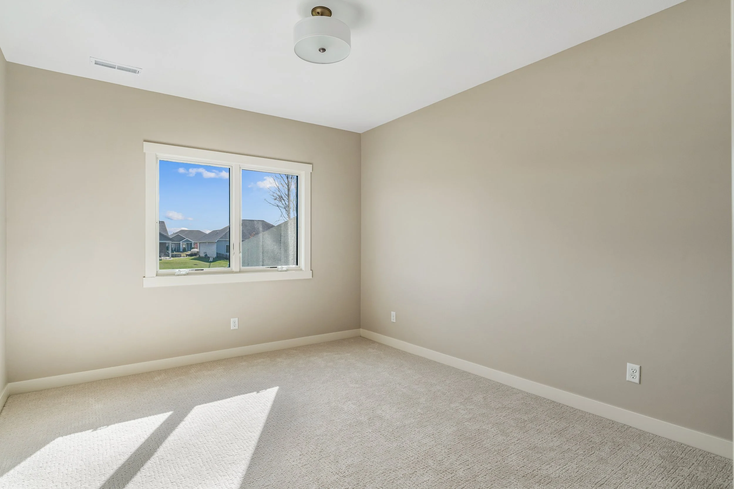 Empty room with beige walls, carpeted floor, a window showing houses outside, and a ceiling light fixture.