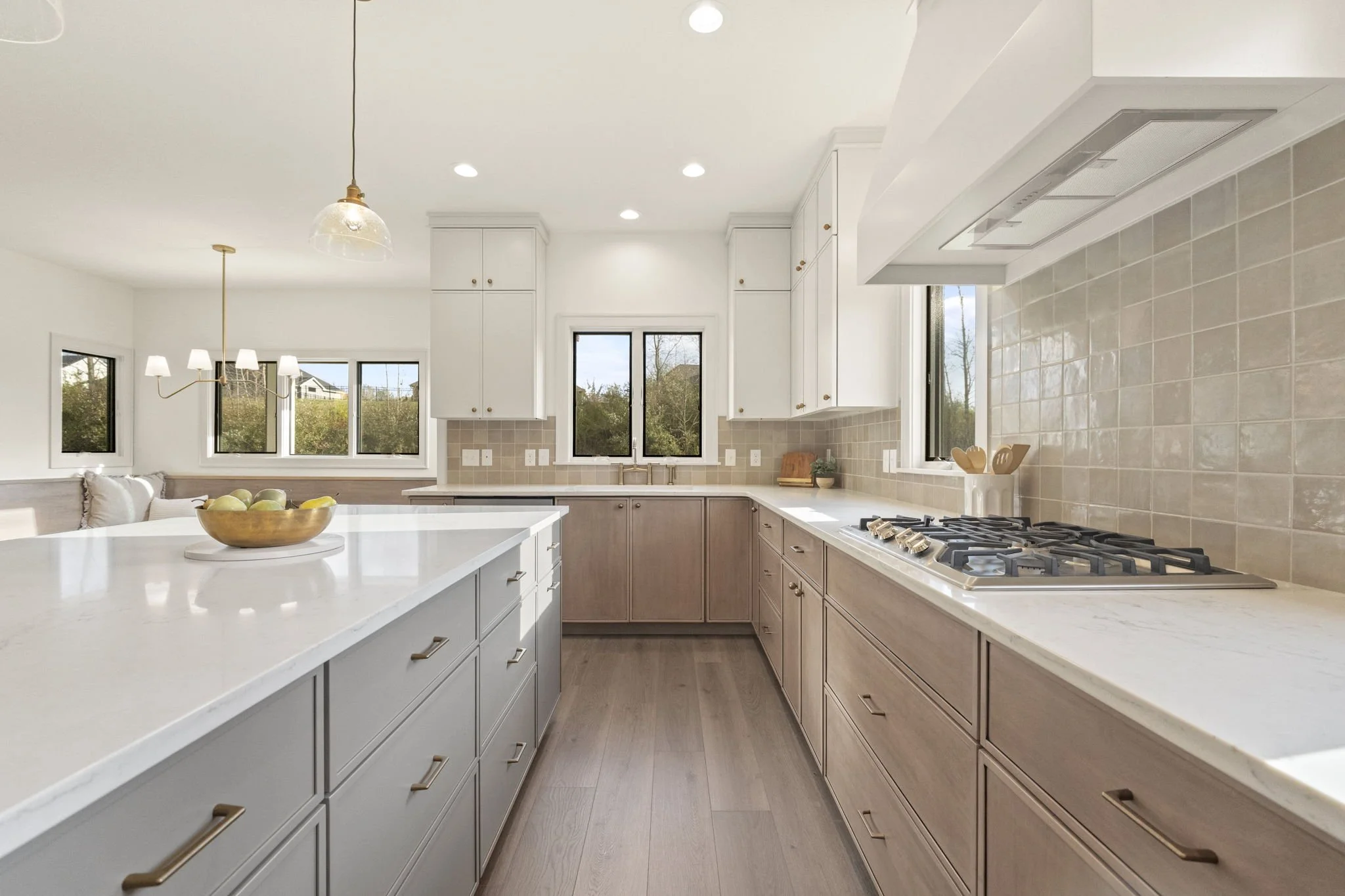 Modern kitchen with white and light wood cabinets, marble countertops, beige tile backsplash, gas stove, and window views of greenery.