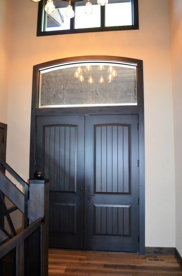 Double black wooden doors with a glass transom window above, inside a modern home with light-colored walls, a staircase to the left, and a wooden floor.