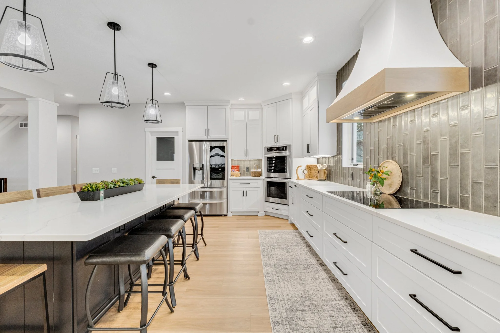 Modern white kitchen with a large island, black bar stools, stainless steel appliances, and a gray tiled backsplash underneath a white range hood.