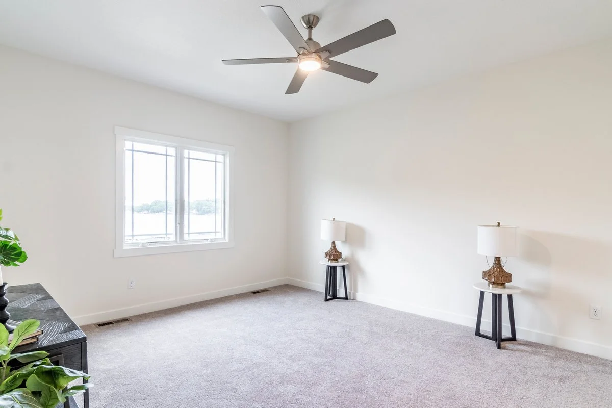 Empty living room with white walls, a large window, a ceiling fan with lights, two side tables with lamps, a black table, and a potted plant.
