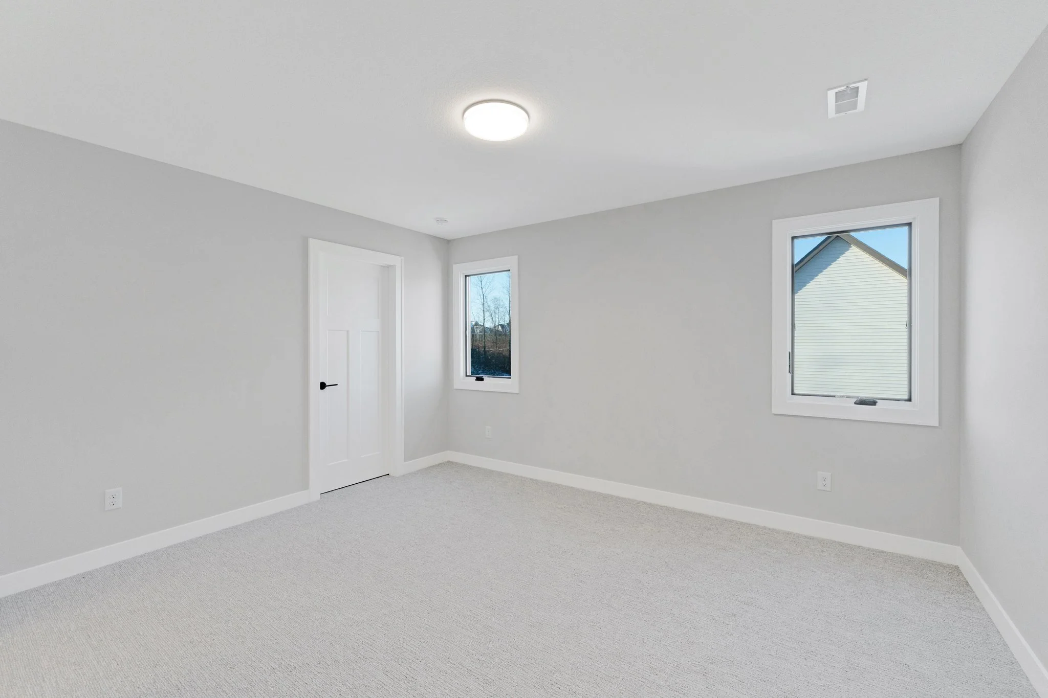 Empty room with white walls, two windows, a closed door, and a ceiling light fixture.