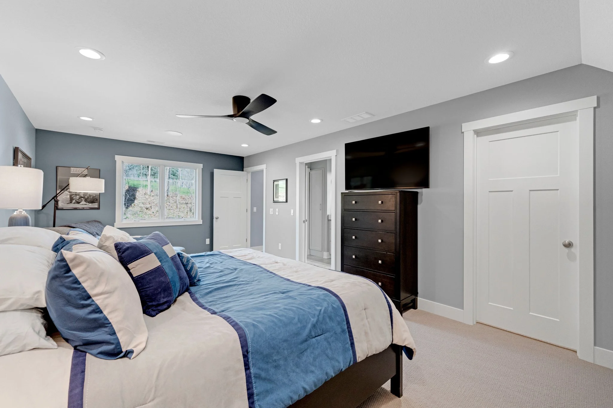 A bedroom with a bed, blue and white bedding, multiple pillows, a window showing trees and hillside outside, a black dresser with a TV on top, white doors, and a ceiling fan with black blades.