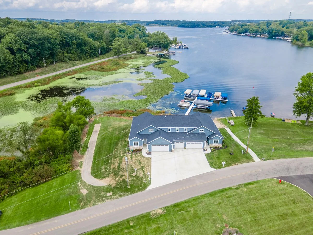 Aerial view of a lakeside house with a large driveway, surrounded by green lawns, trees, and a boat dock with several boats, overlooking a lake with green aquatic vegetation and a forested shoreline.