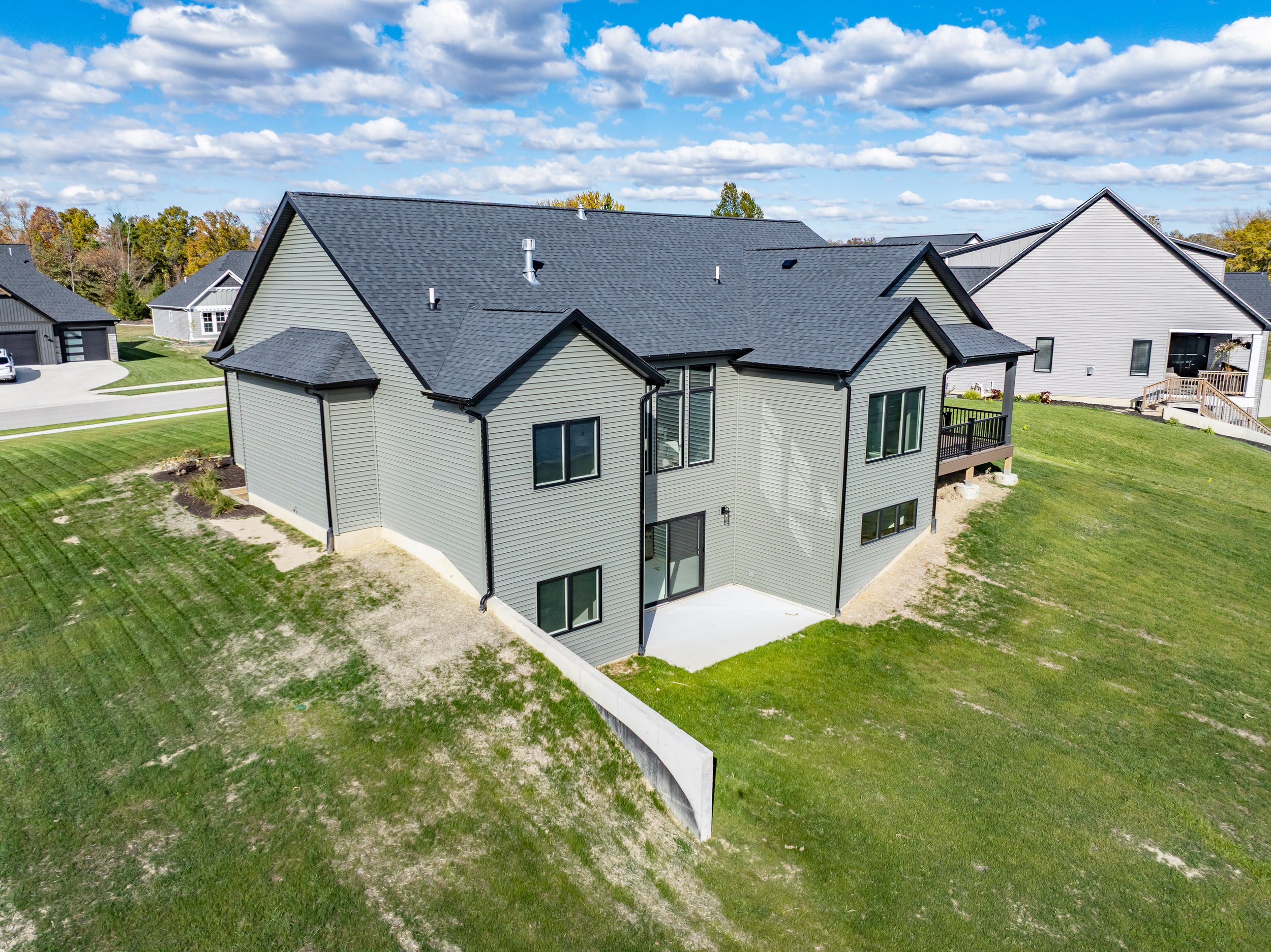 A modern two-story house with light grey siding, black window frames, and a dark roof on a spacious grassy lot with neighboring houses nearby, under a partly cloudy sky.