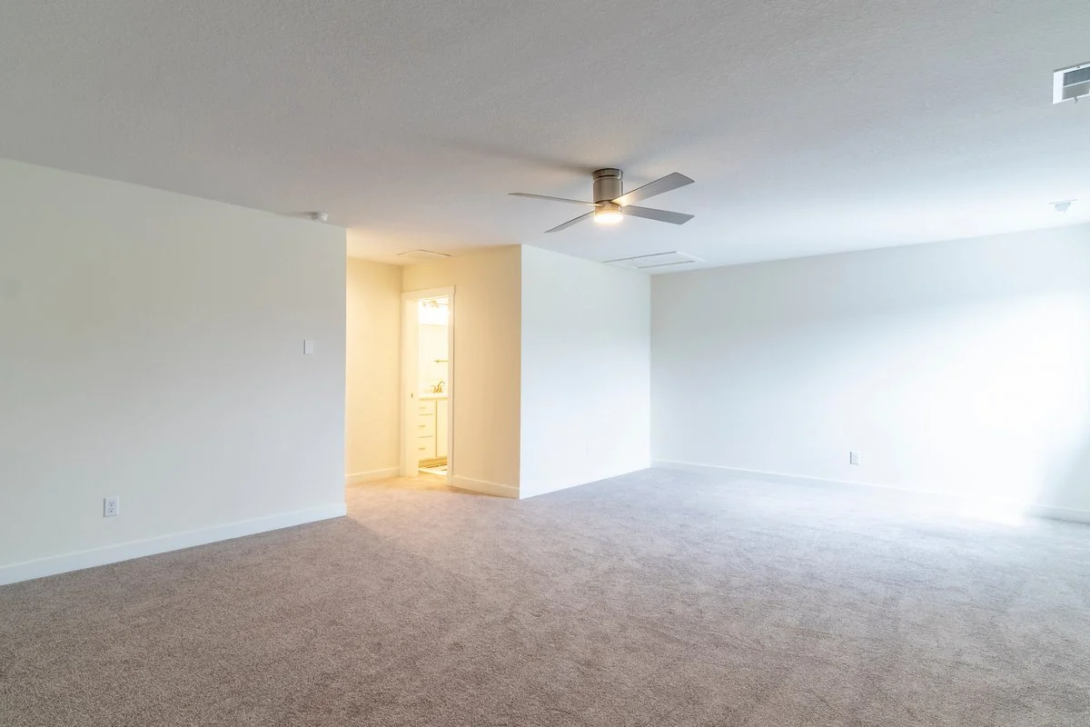 Empty living room with beige carpet, white walls, ceiling fan, and natural light from windows.