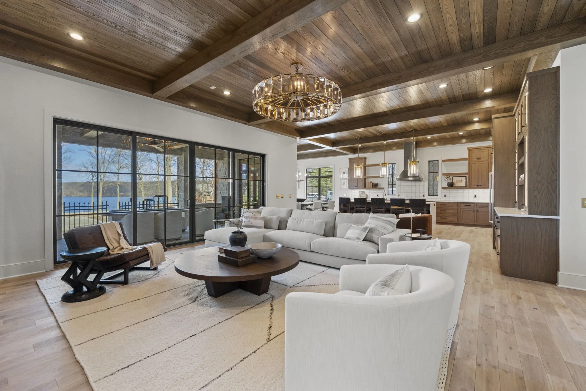 Living room with white sectional sofa, two white armchairs, a round wooden coffee table, and large glass sliding doors overlooking a lake. The room has wooden ceiling beams and hardwood flooring, with a view of a kitchen area in the background.