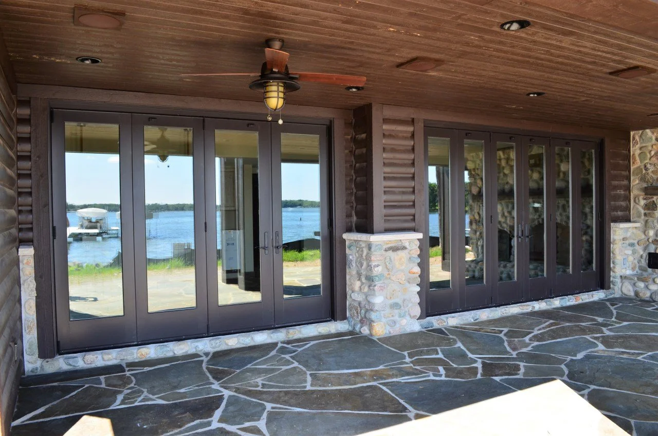A porch area with stone and wood details, featuring large glass double doors and a ceiling fan, overlooking a lake with boats.