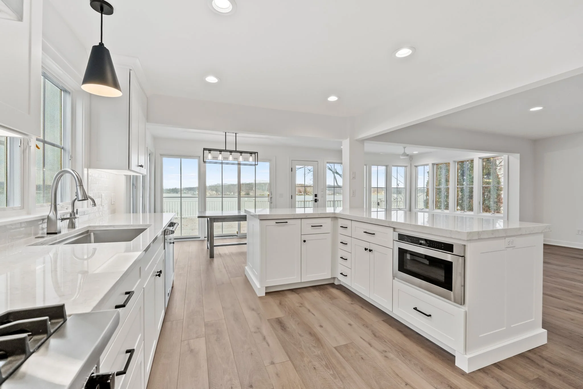 Bright white kitchen with wooden flooring, white cabinets, and large windows overlooking a scenic landscape. Features an island, built-in stove, oven, and modern lighting fixtures.