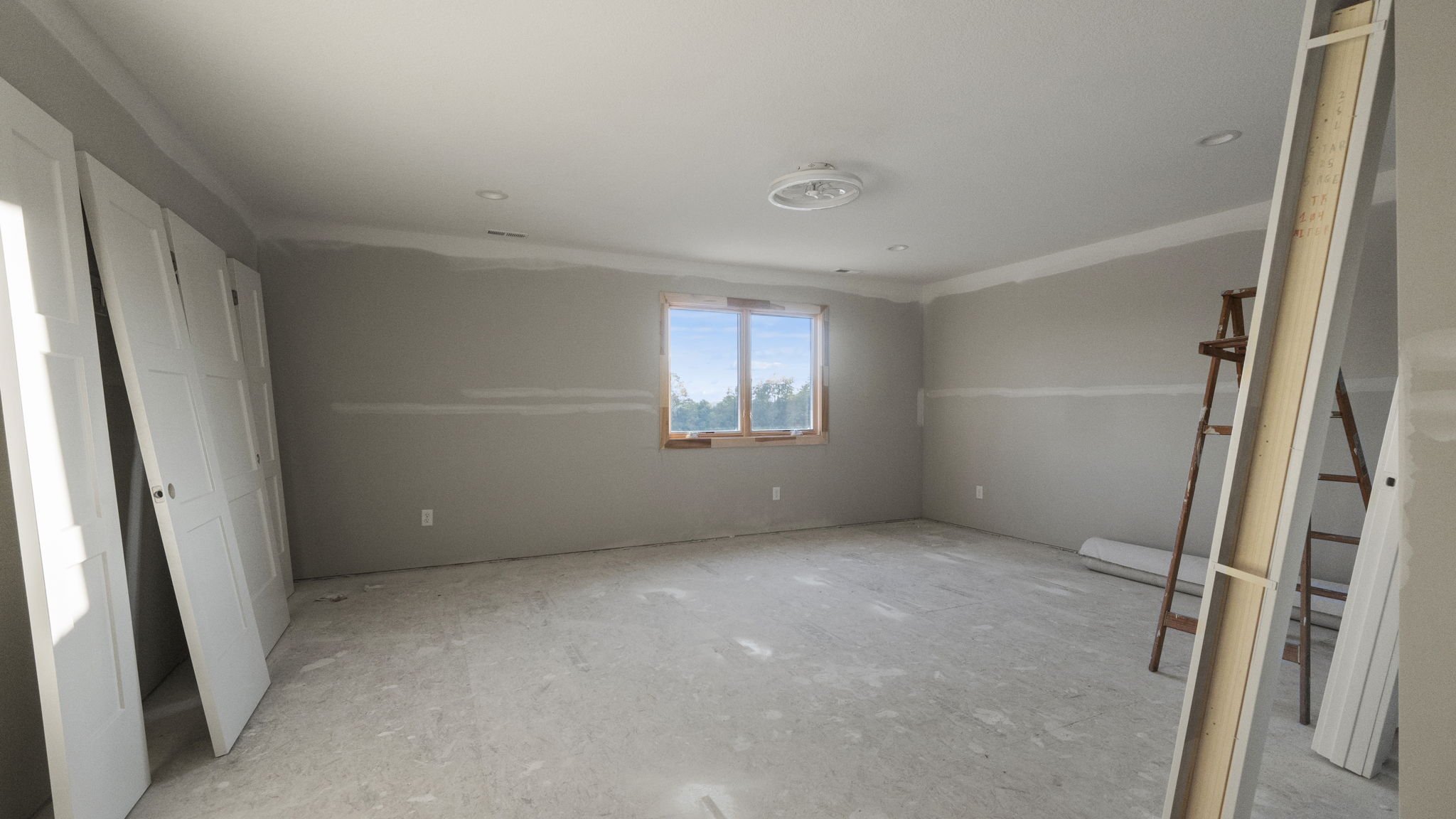 Unfinished empty room with drywall walls, a window, and construction materials including a ladder and doors leaning against the wall.