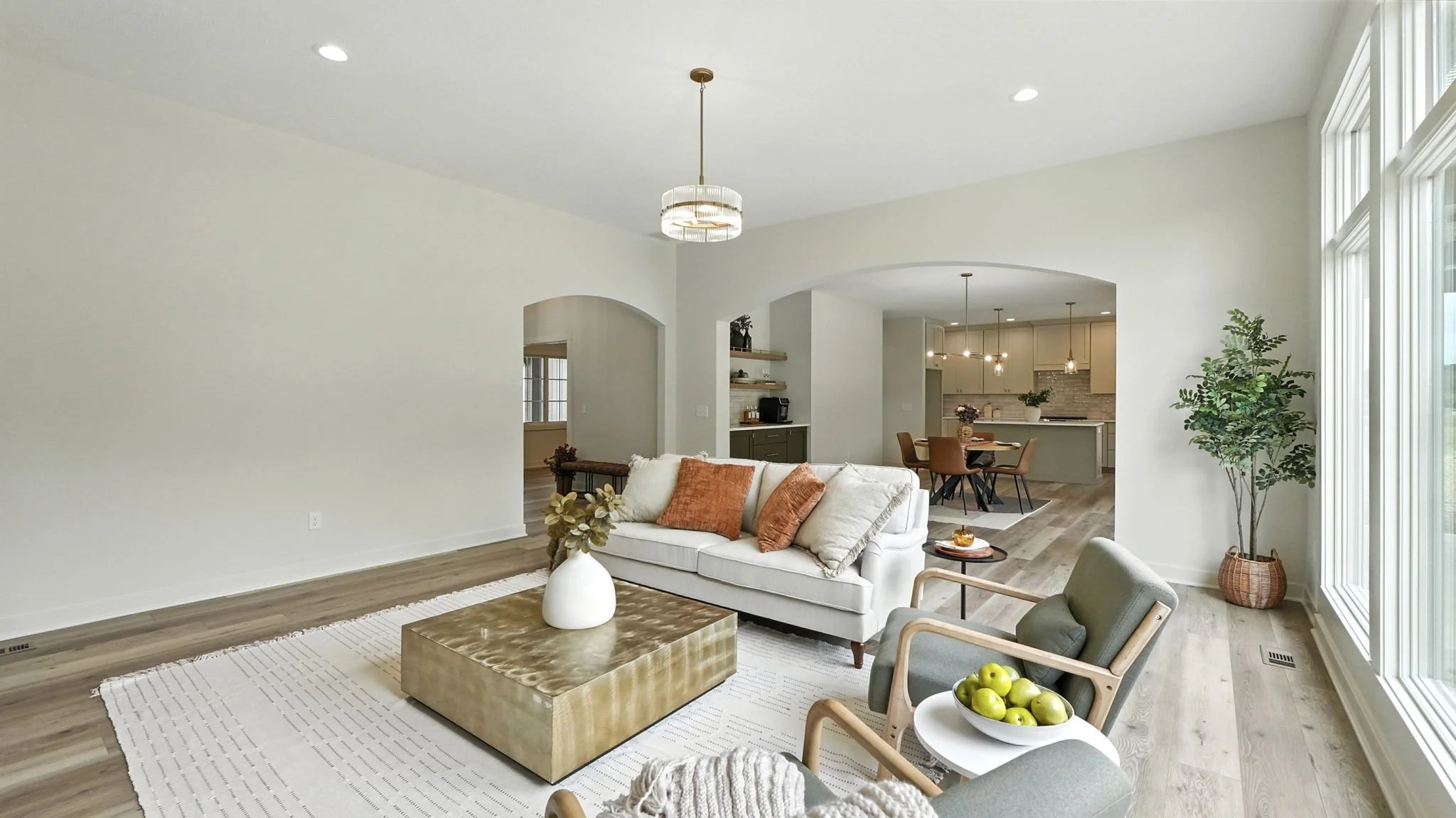 Living room with white sofa, green and orange throw pillows, beige armchair with bowl of green apples, large window, potted plant, view into dining and kitchen area with wooden chairs, pendant lights, and kitchen island.