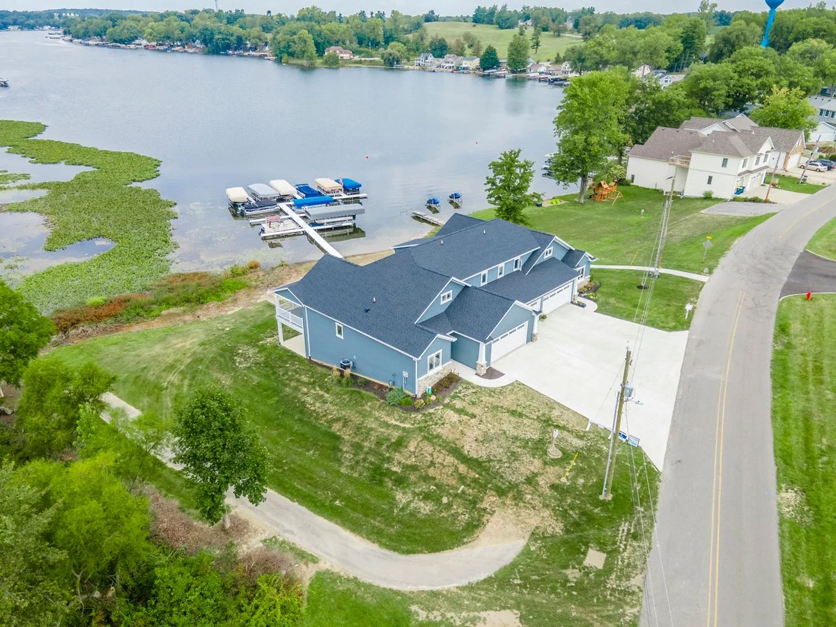 Aerial view of a house by a lake with boat docks, green lawns, trees, and neighboring houses.