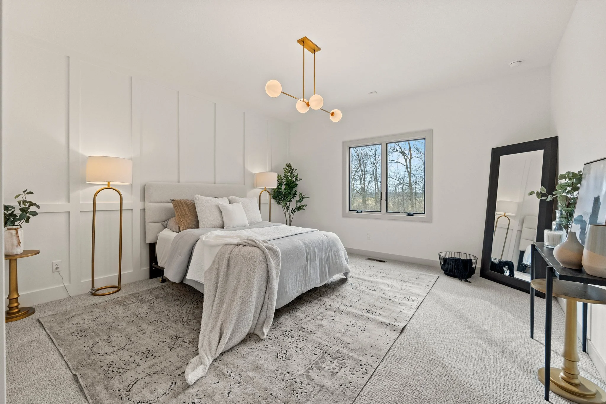 A modern bedroom with white walls, a bed with white bedding and multiple pillows, two standing lamps with white shades and gold stands on each side of the bed, a large black-framed mirror leaning against the wall, a window overlooking trees, a small 