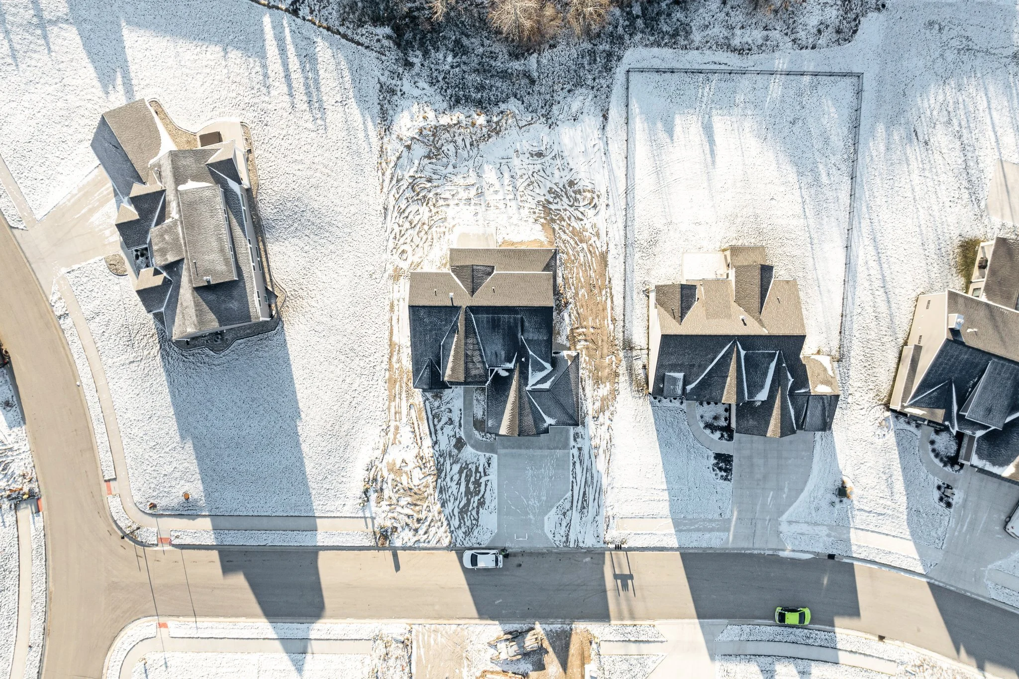 An aerial view of a residential neighborhood in winter, showing four houses with snow-covered roofs, a paved street, and parked cars.