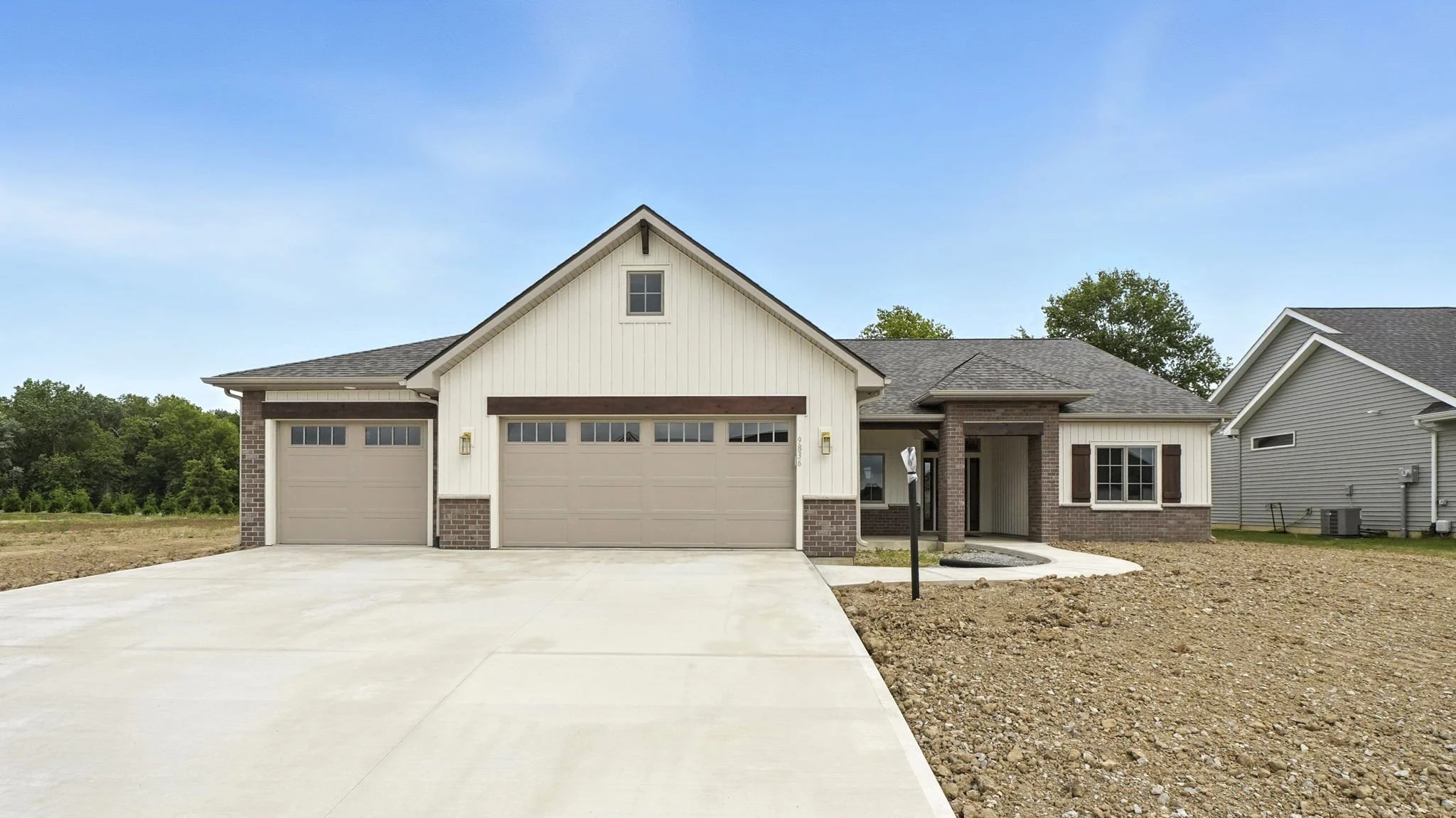 Newly built suburban house with three-car garage, beige doors, brick accents, white siding, and a concrete driveway, with partly landscaped yard and neighboring homes visible.