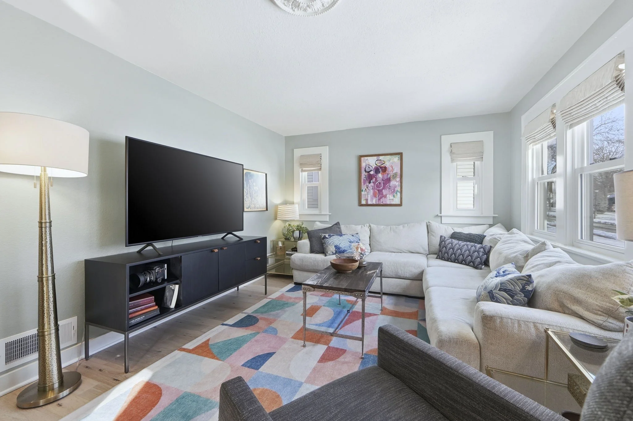 Living room with a large window, white sectional sofa with patterned pillows, a dark coffee table, a blue TV stand with a large flat-screen TV, colorful geometric area rug, night stand with a lamp, and artwork on the wall.