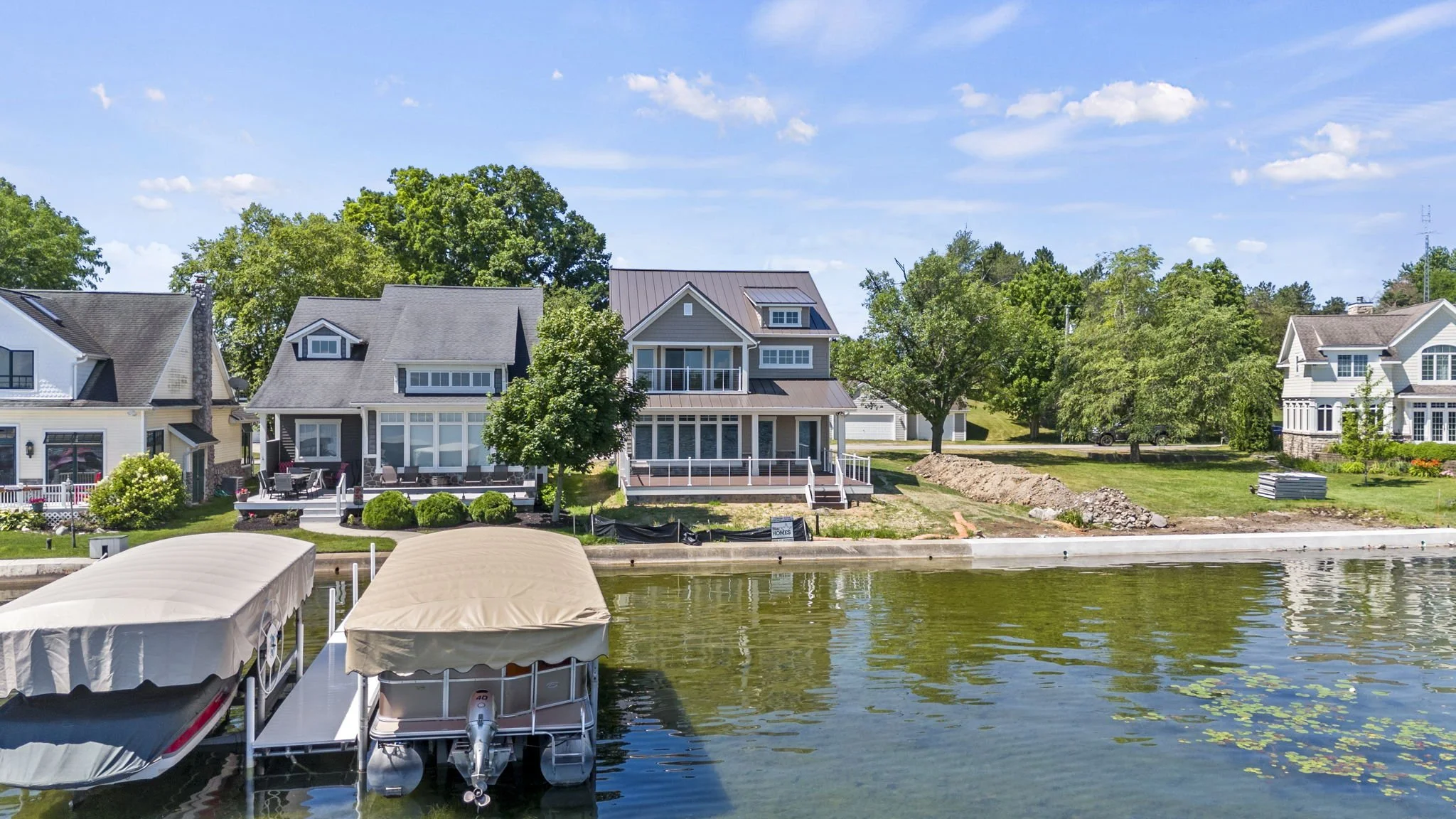Residential houses along a lakeside, with a dock and boats in the foreground, green trees, and blue sky with clouds in the background.