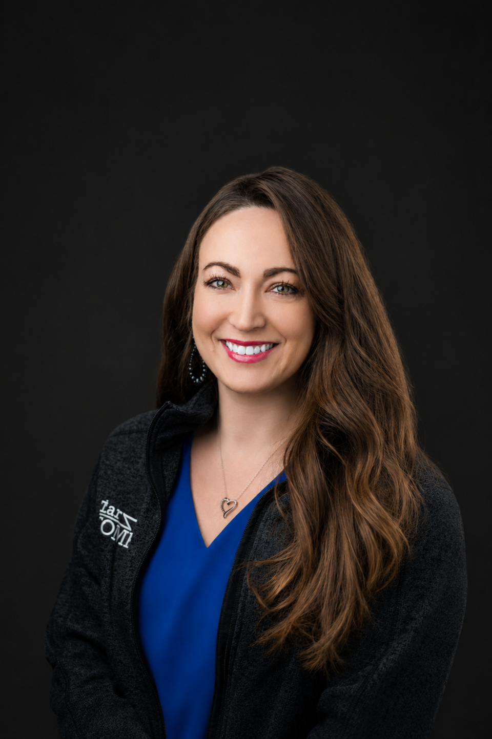 A woman with long brown hair smiling, wearing a dark jacket, posing with her arms crossed against a dark background.