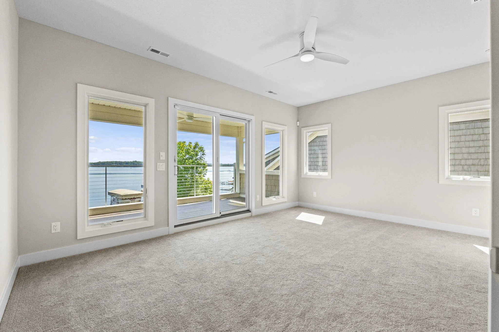 Empty living room with large windows and a sliding glass door overlooking a water view and a balcony, beige carpet, white ceiling with a fan, and white walls.