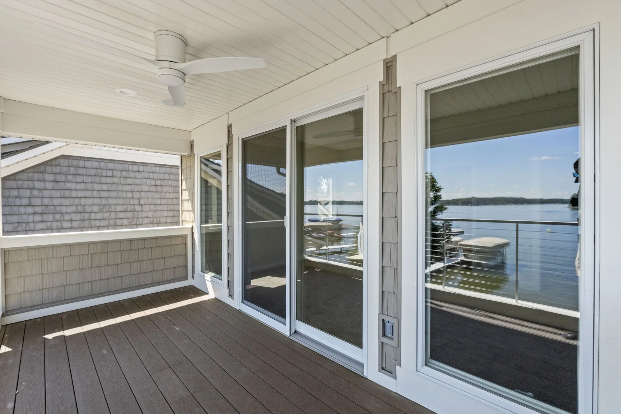 View of a deck with glass sliding doors and large windows, overlooking a body of water with boats and trees in the background, under a covered porch with a ceiling fan.