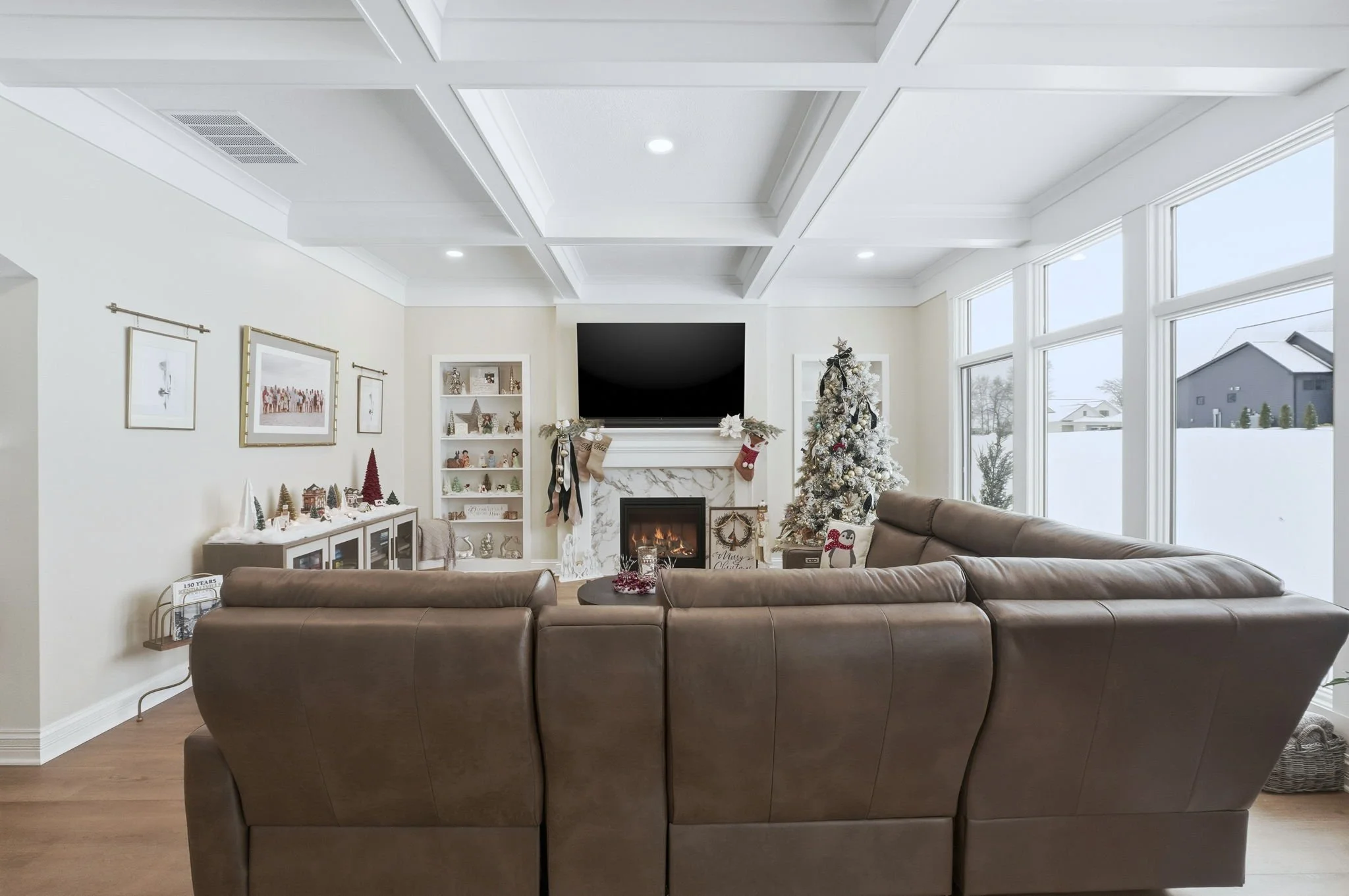 Living room with a brown sectional sofa, decorated Christmas tree, fireplace, wall-mounted TV, and large windows showing snowy outdoor scene.