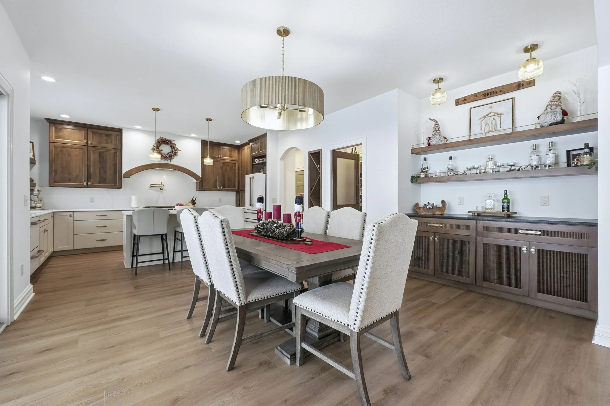 Open concept kitchen and dining area decorated for Christmas with a large wooden dining table, upholstered chairs, wooden cabinets, and holiday decorations including garland and Santa figurines.