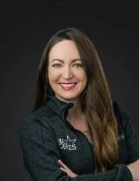 A woman with long brown hair smiling, wearing a dark jacket, posing with her arms crossed against a dark background.