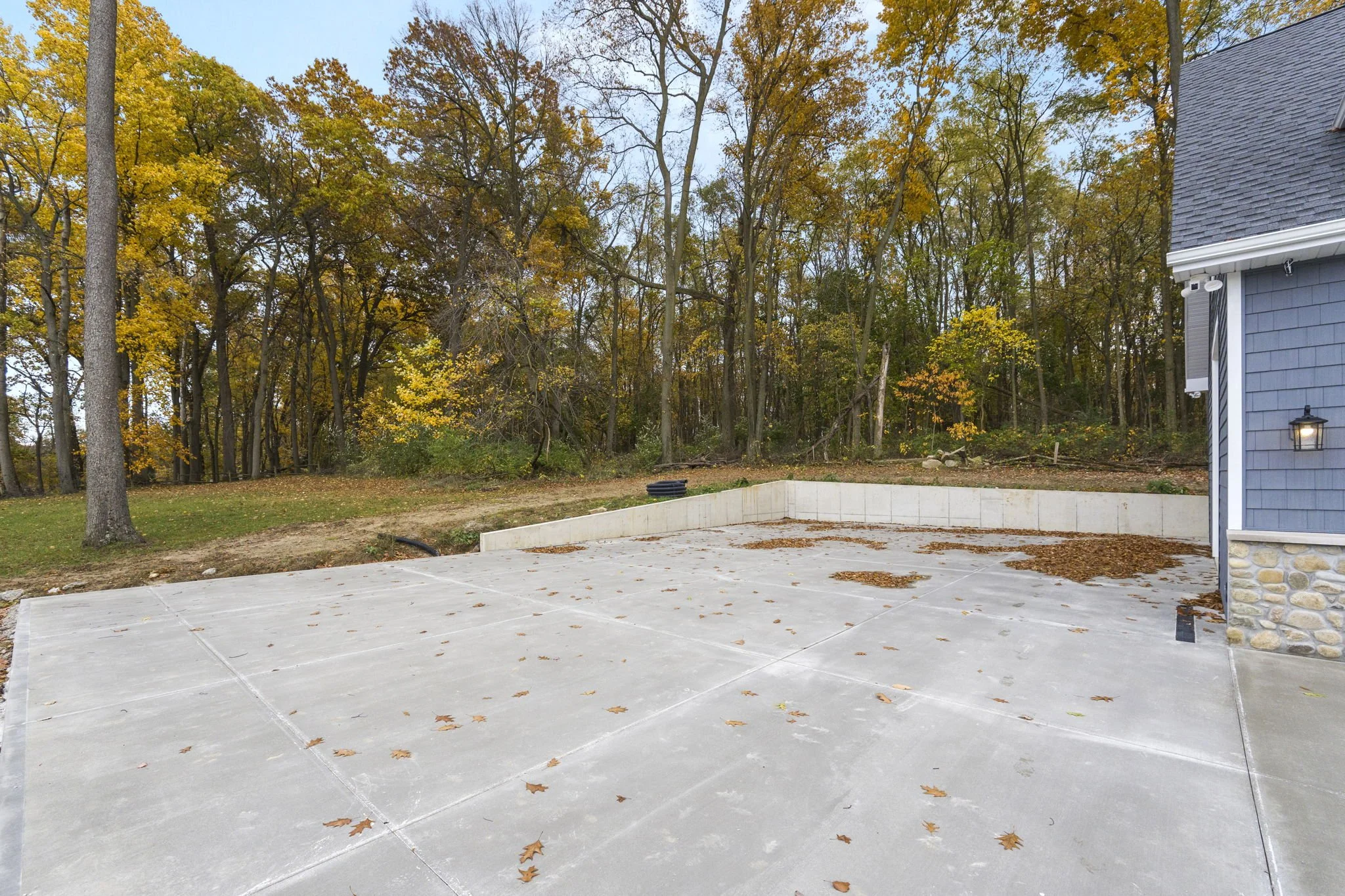 Empty concrete patio with fallen leaves, adjacent to a house with gray siding, and a wooded area with trees showing fall foliage in the background.