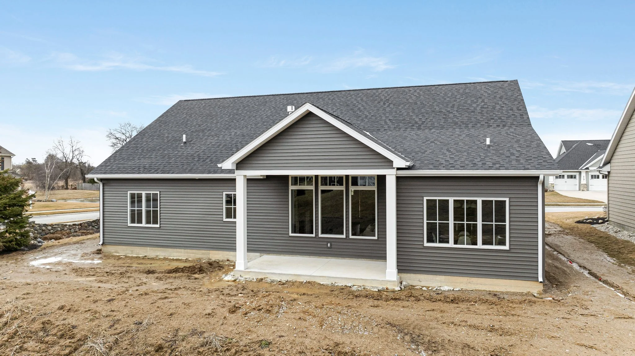 Newly constructed house with gray siding, black shingle roof, and a small covered patio at the back, surrounded by a dirt yard and neighboring houses in the background.