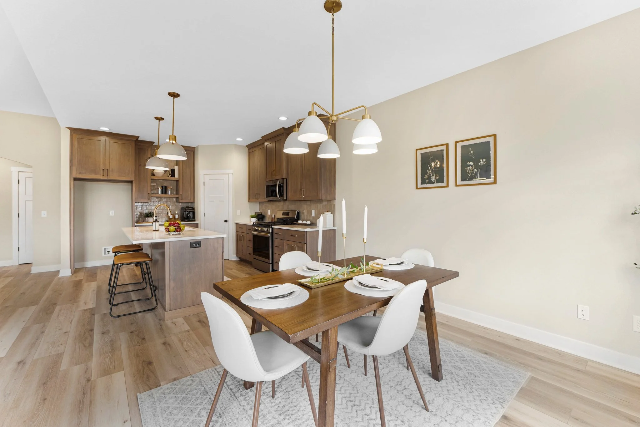 Dining area with a wooden table set with white plates and candles, surrounded by white chairs, next to an open kitchen with wooden cabinets, stainless steel appliances, and an island with stools.