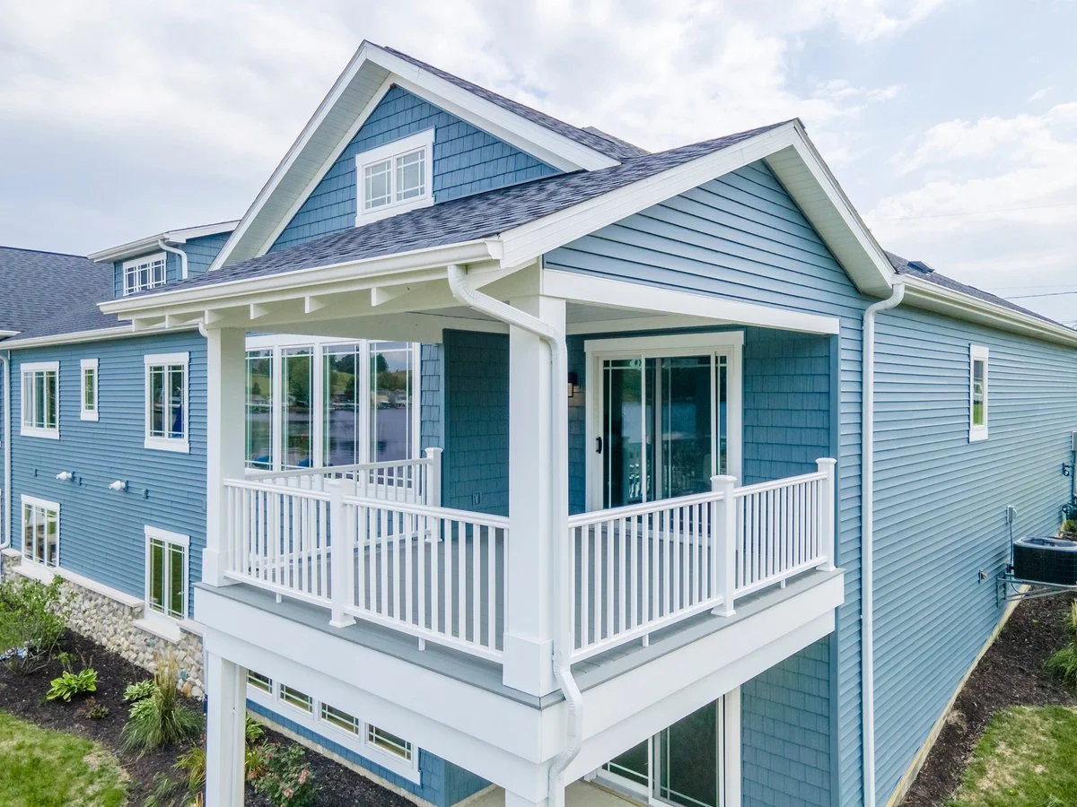 A two-story blue house with white trim and a balcony with a white railing. The house has multiple windows and a gable roof.