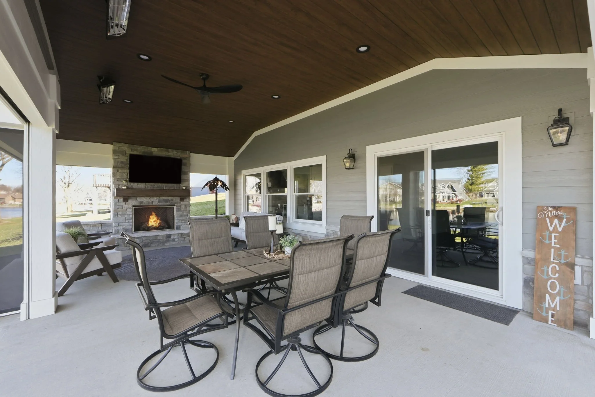 Covered outdoor patio area with a dining table and eight chairs, a fireplace with a TV above, and a sliding glass door leading to the interior house, decorated with a welcome sign and nautical-themed decor.