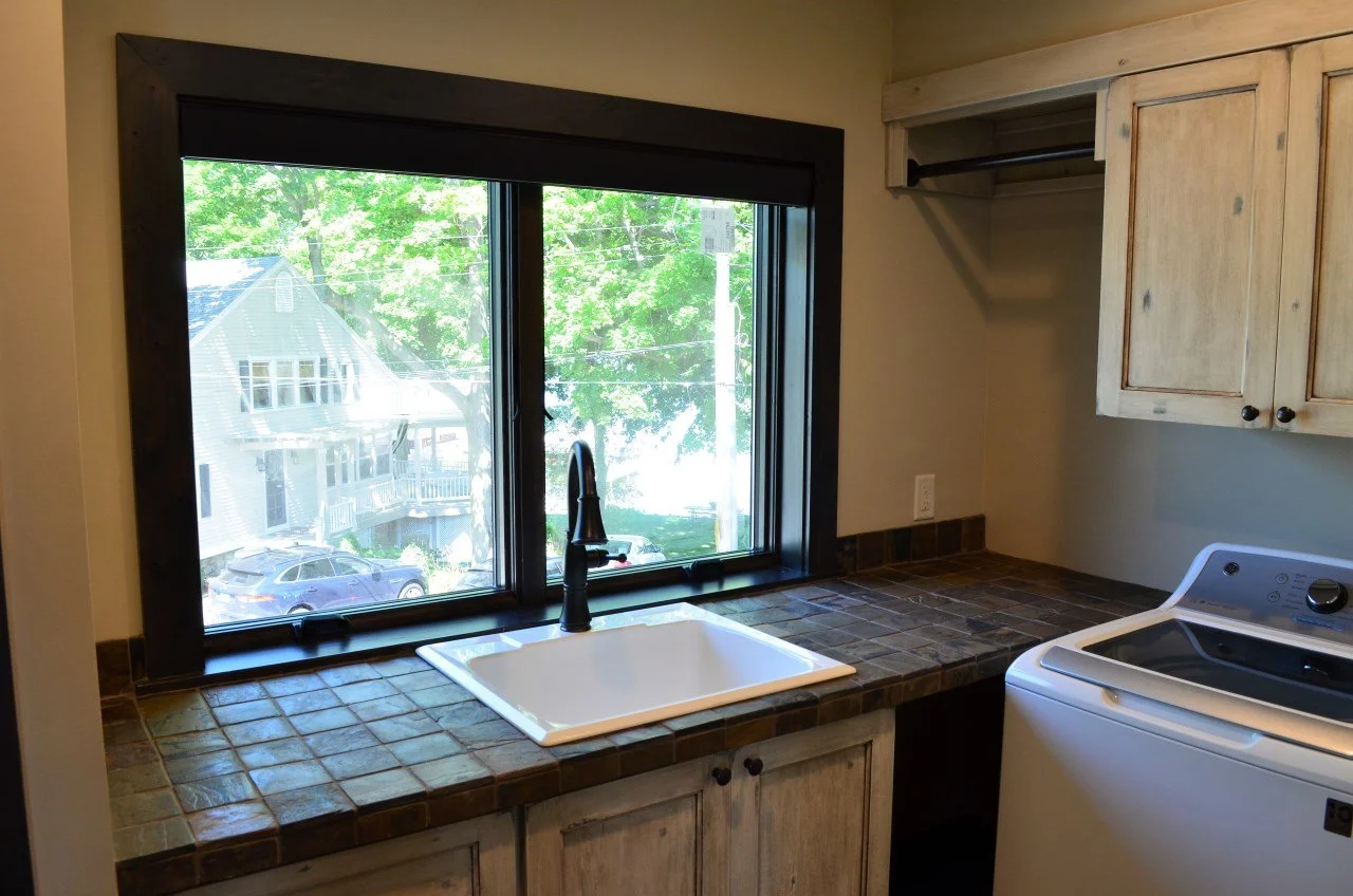 Kitchen with a large window, a white farmhouse sink, a black faucet, tile countertop, upper cabinets, and a washing machine.