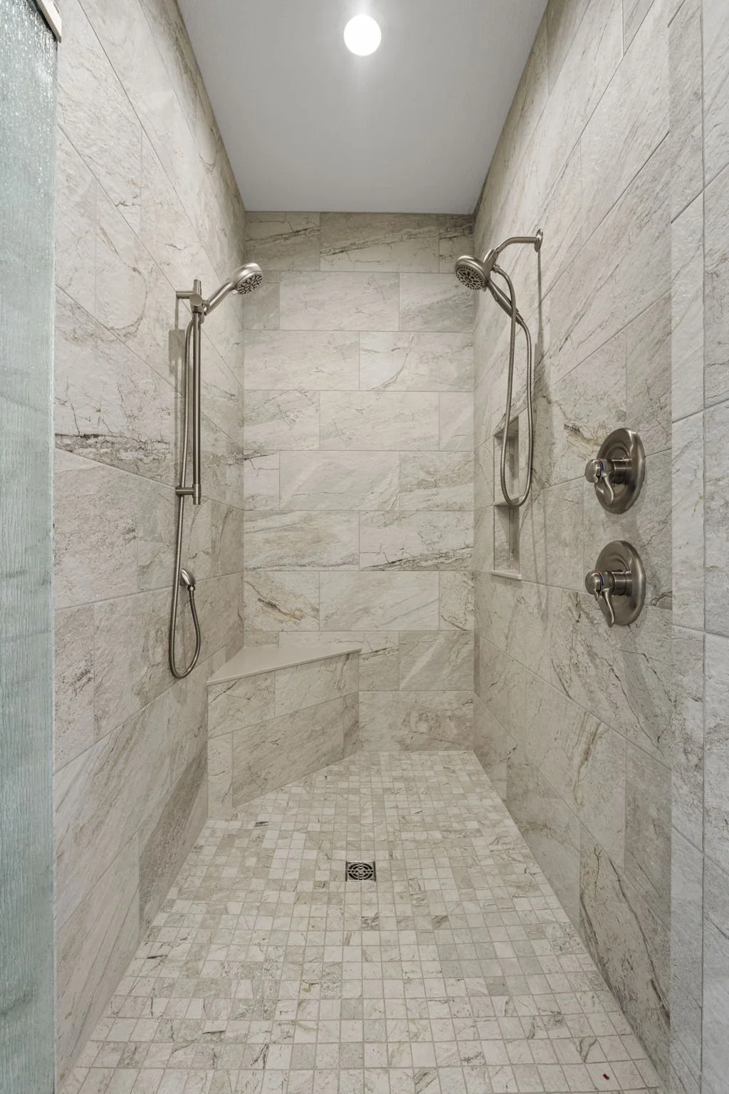 A clean, modern walk-in shower with beige marble tiles, two showerheads, and two controls on the right wall.