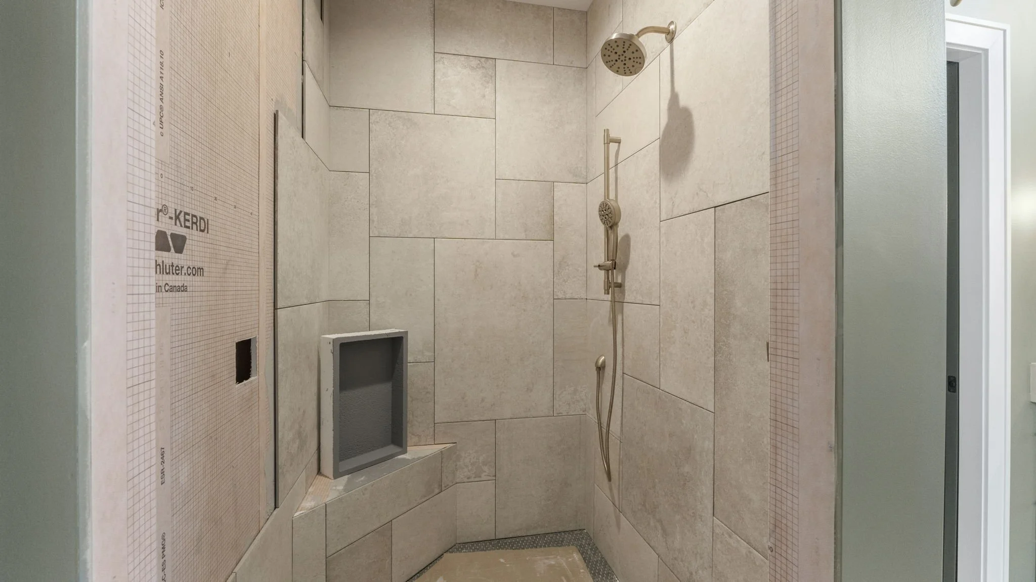A shower area under construction with beige tiles on the walls and a built-in seat. There is a showerhead and a handheld shower attachment.