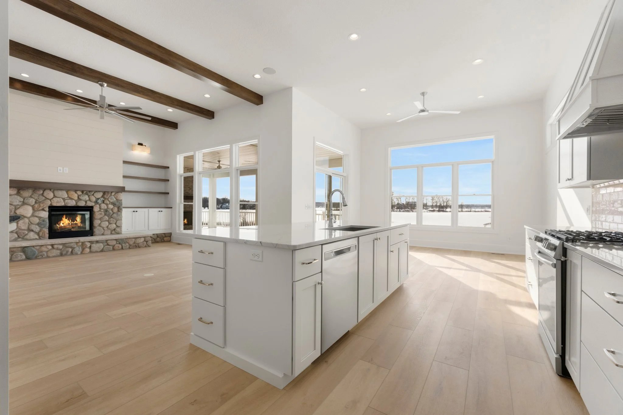 Bright open kitchen and living area with large windows, white cabinets, a stone fireplace, and wooden beams on the ceiling.