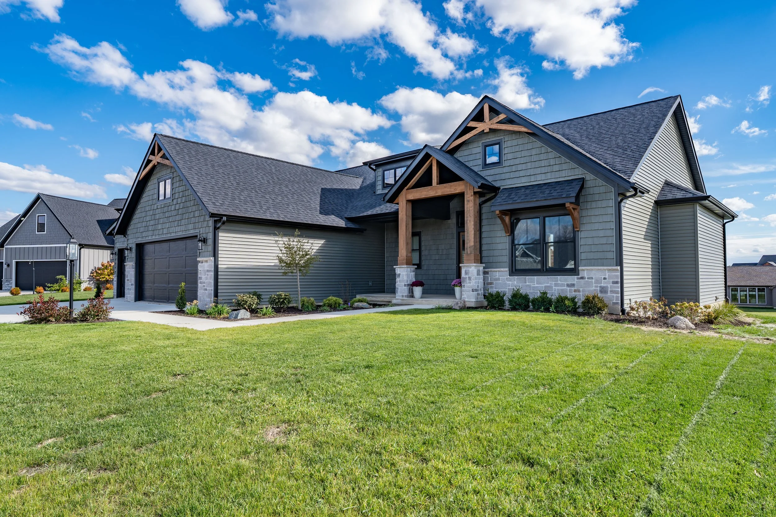 A modern, two-story house with gray siding, black roof, and a front porch with wooden beams and stone pillars, surrounded by a well-maintained lawn and landscaping, under a partly cloudy blue sky.