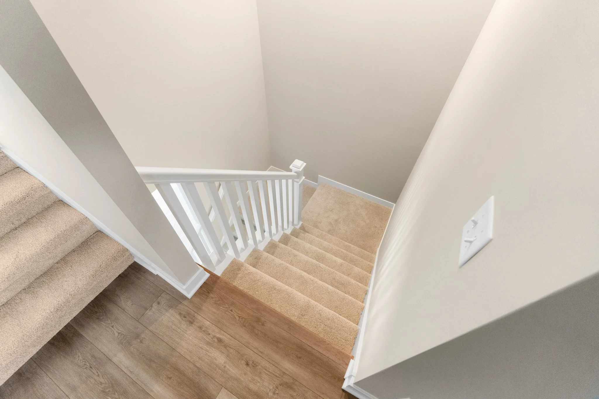 Top-down view of a staircase with beige carpeted steps, white handrail, and beige walls, leading down from a hardwood floor.