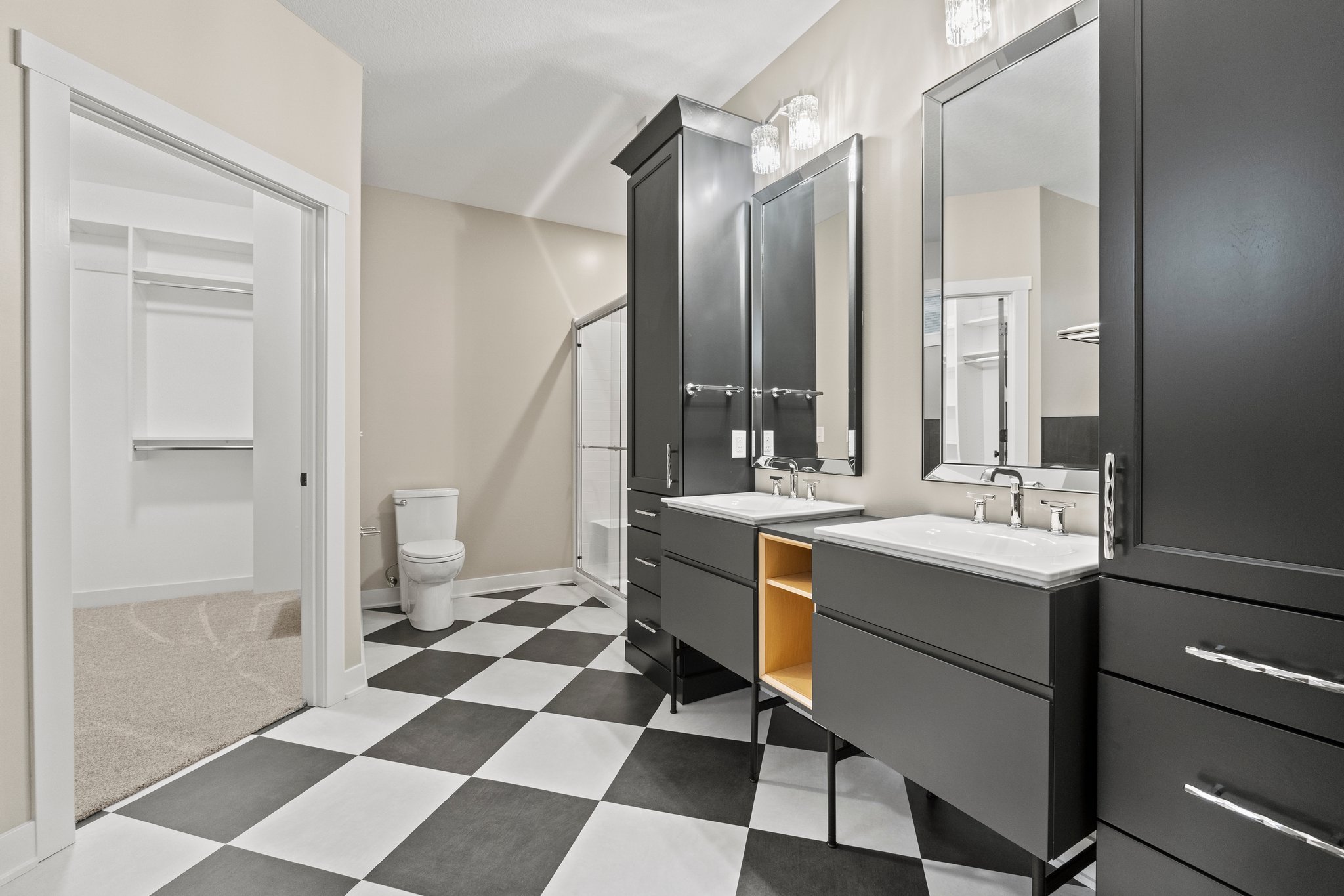 The image shows a modern bathroom with black and white checkerboard floor tiles, a double sink vanity with black cabinets and white sinks, mirrors above each sink, a black tall storage cabinet, and a separate room with a toilet and an open closet spa