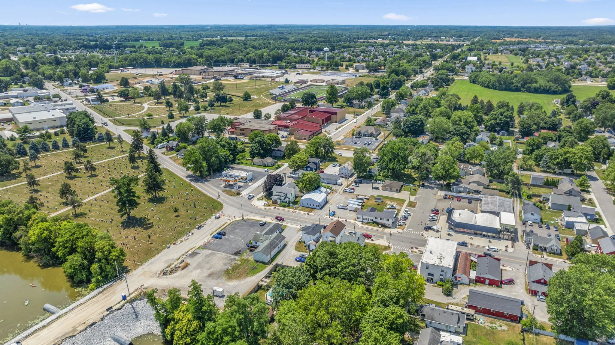 Aerial view of a small town with residential houses, commercial buildings, a parking lot, a river, and green spaces under a clear blue sky.
