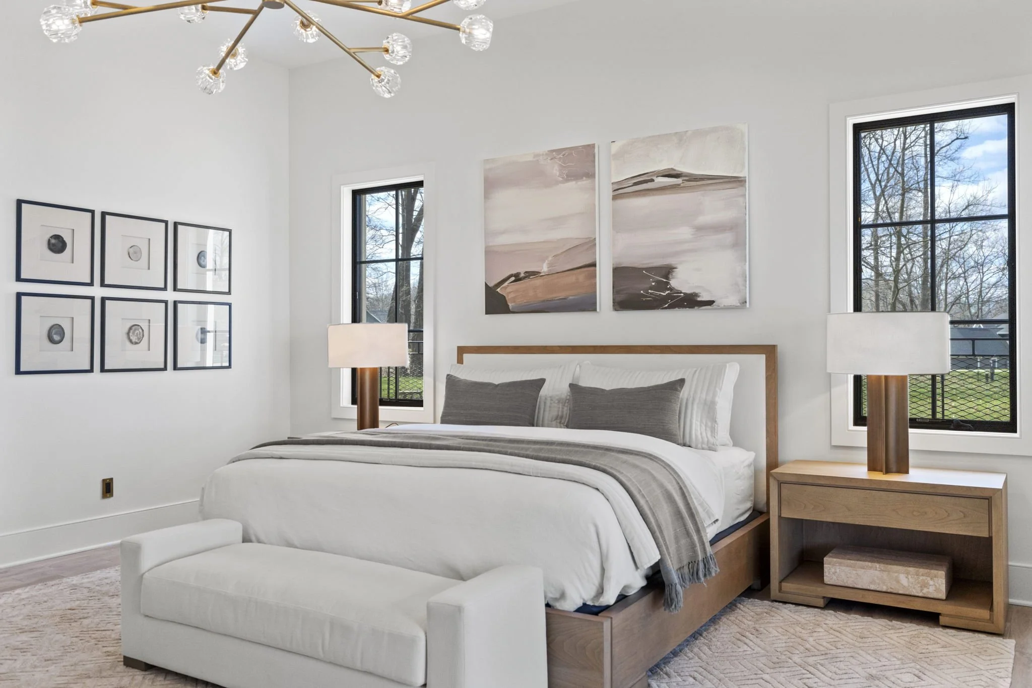 A modern bedroom with a white bed, gray pillows, and a gray throw blanket. There are two wooden nightstands with table lamps on either side of the bed. The room has two large windows revealing trees outside. Artwork adorns the walls, including two ab