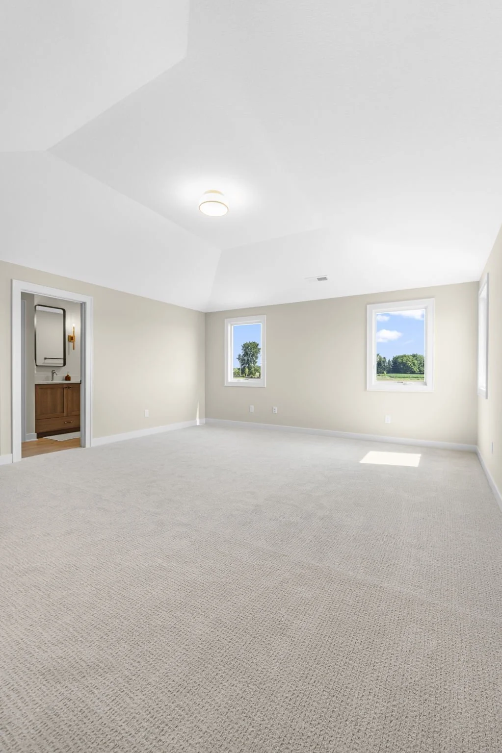 Empty room with beige walls, three windows showing outdoors, light-colored carpet, ceiling light fixture, and a doorway leading to a bathroom with wooden vanity and mirror.