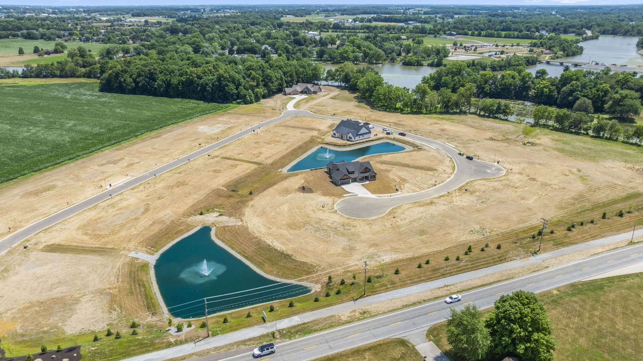 Aerial view of a large construction site with multiple newly built houses, lakes with fountains, and winding roads in a semi-rural area surrounded by green fields, trees, and a body of water in the background.
