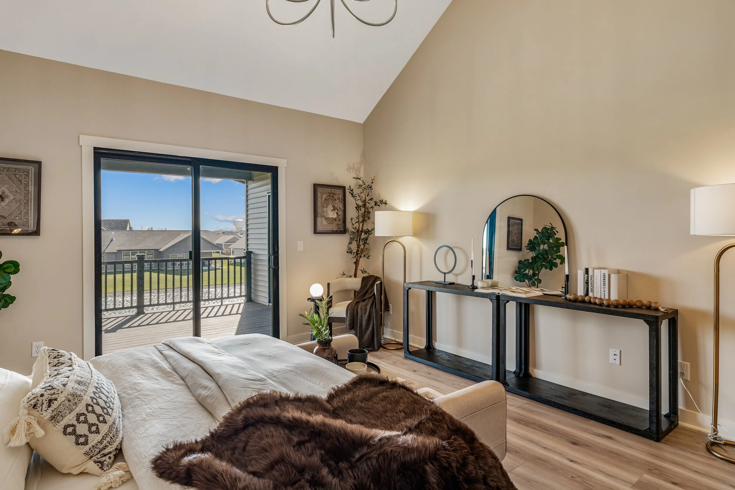 A cozy bedroom with a sliding glass door opening to a balcony, overlooking houses and a blue sky. The room features a bed with a brown fur throw, a beige armchair, a black console table with decorative items, and floor lamps. There are framed artwork