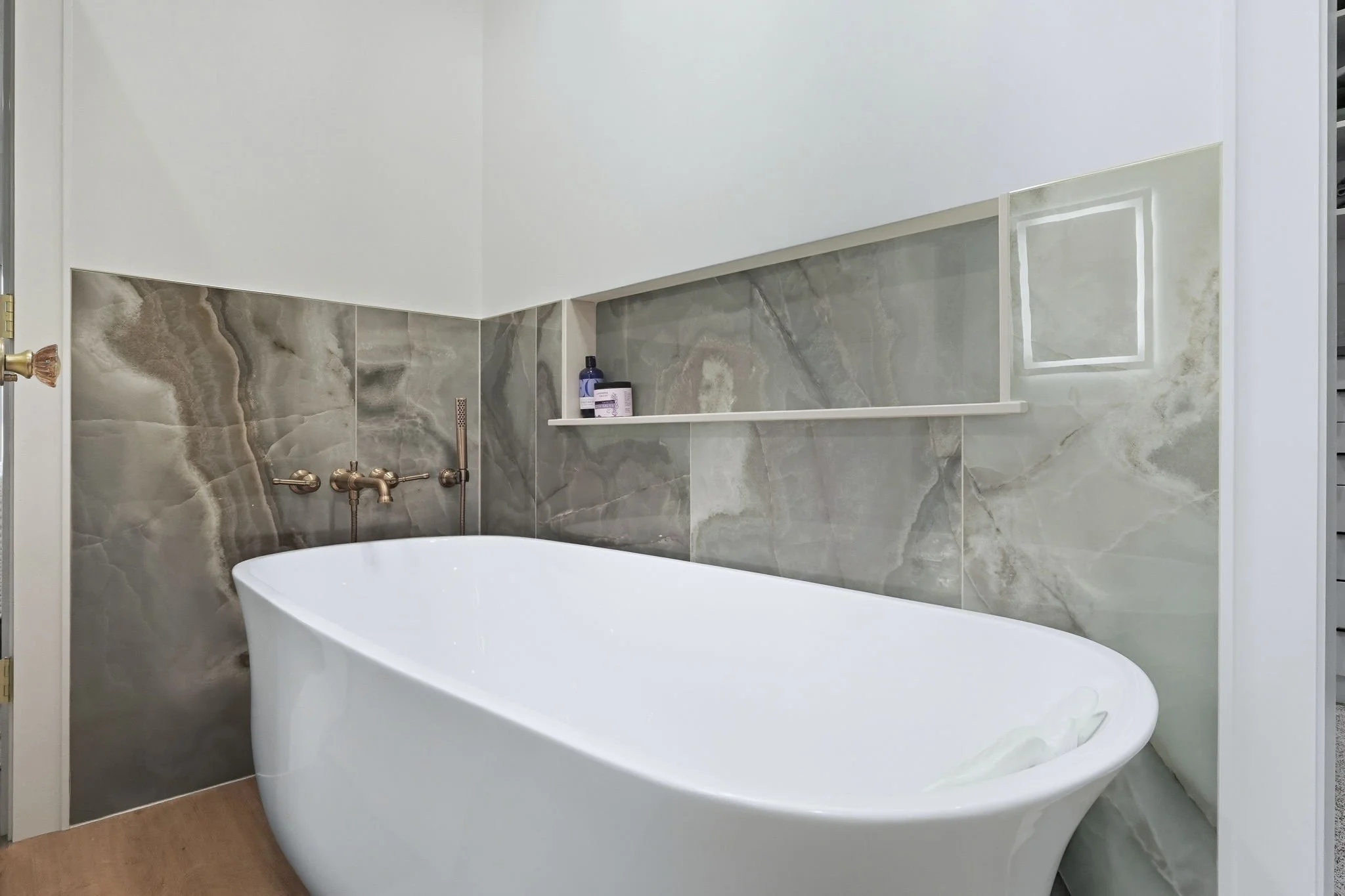 Modern bathroom with a white bathtub, gray marble tiles on the walls, and a built-in shelf with toiletries.