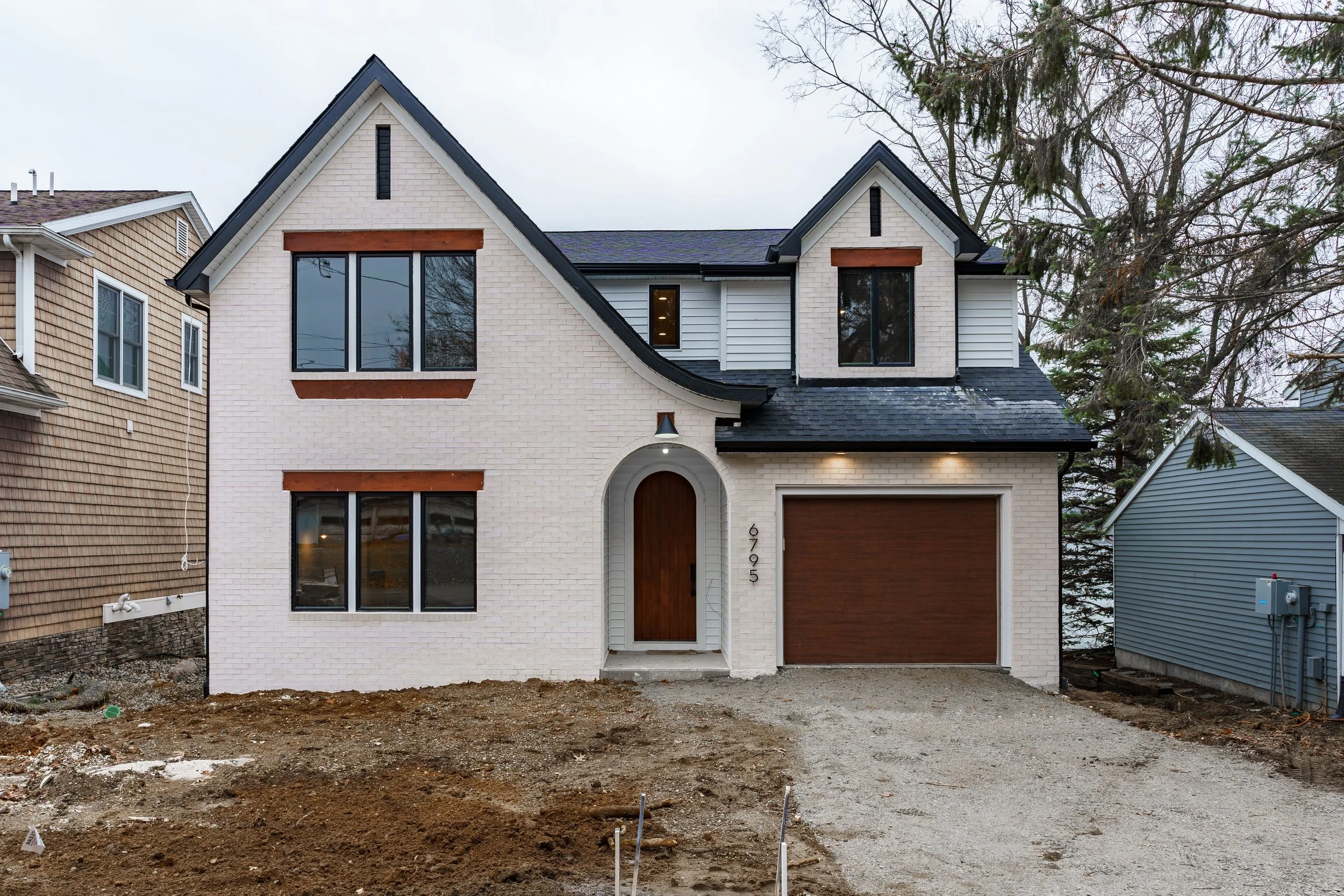 Newly built two-story house with white brick exterior, dark roof, and a front garage with a wooden door, surrounded by neighboring houses and trees, with a dirt driveway in front.
