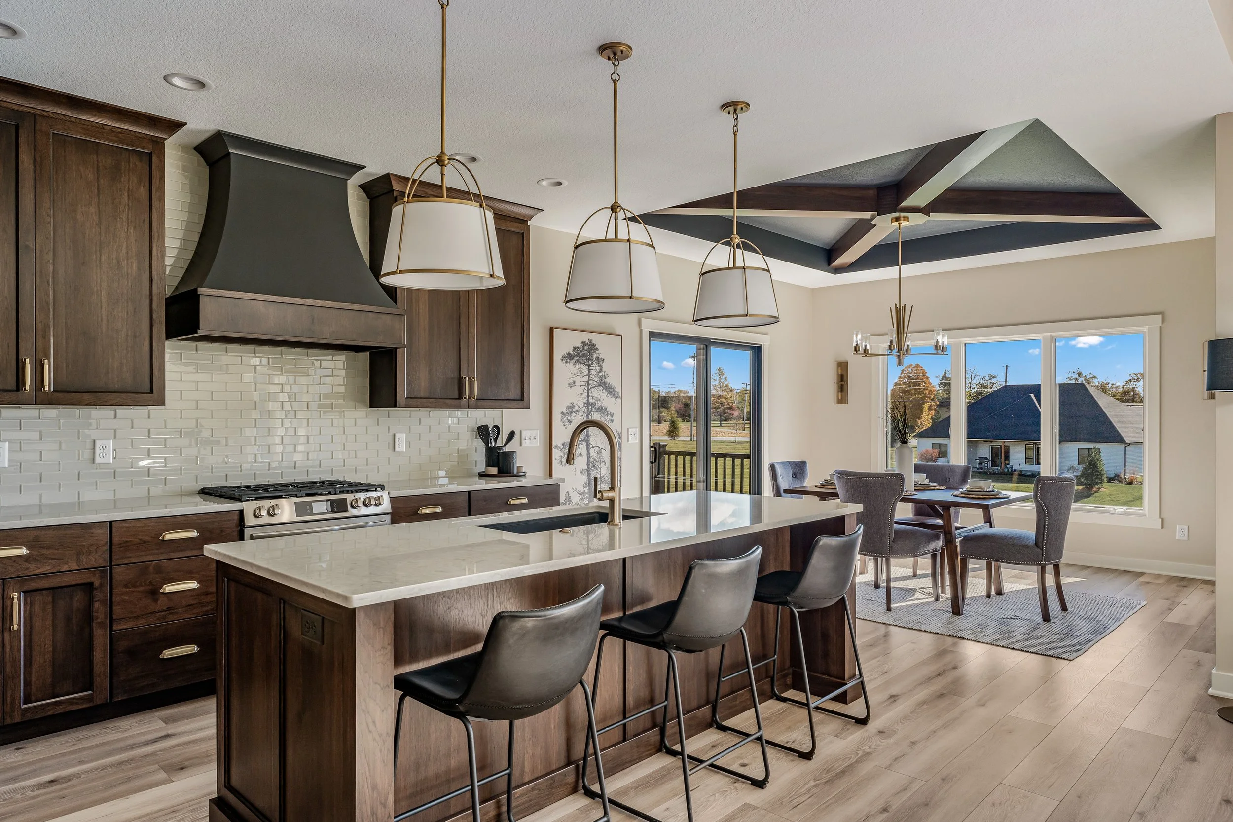 Modern kitchen with dark wood cabinets, a white island with seating, pendant lights, a gas stove, and a dining area with gray chairs and large windows showing a view of a neighborhood.